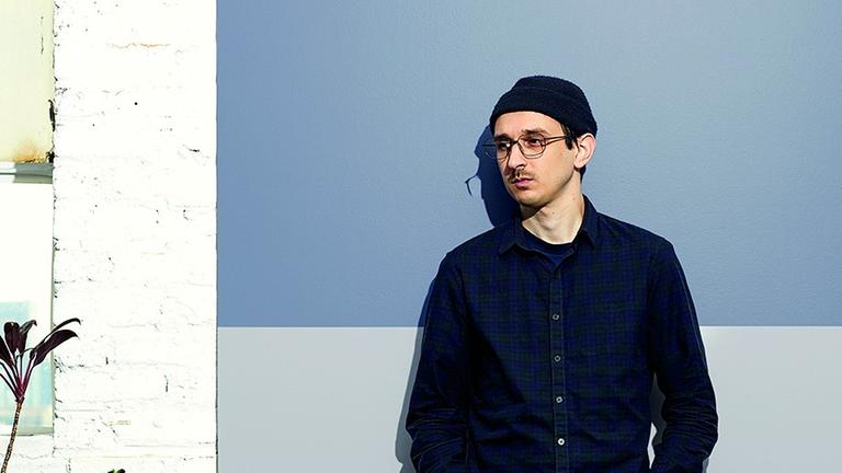 Der Comicautor Nick Drnaso steht, gekleidert mit schwarzer Hose, Pullover und Mütze, vor einer Wand und blick nach rechts unten. Der Comicautor Nick Drnaso steht, gekleidert mit schwarzer Hose, Pullover und Mütze, vor einer Wand und blick nach rechts unten.