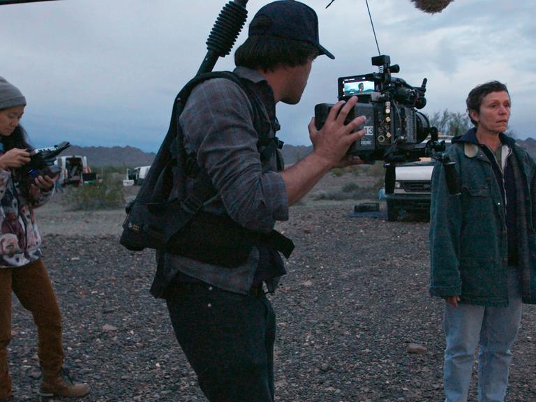 Regisseurin Chloé Zhao, Kameramann Joshua James Richards und Frances McDormand beim Dreh zu "Nomadland". Vor karger Landschaft und bewölktem Himmel.