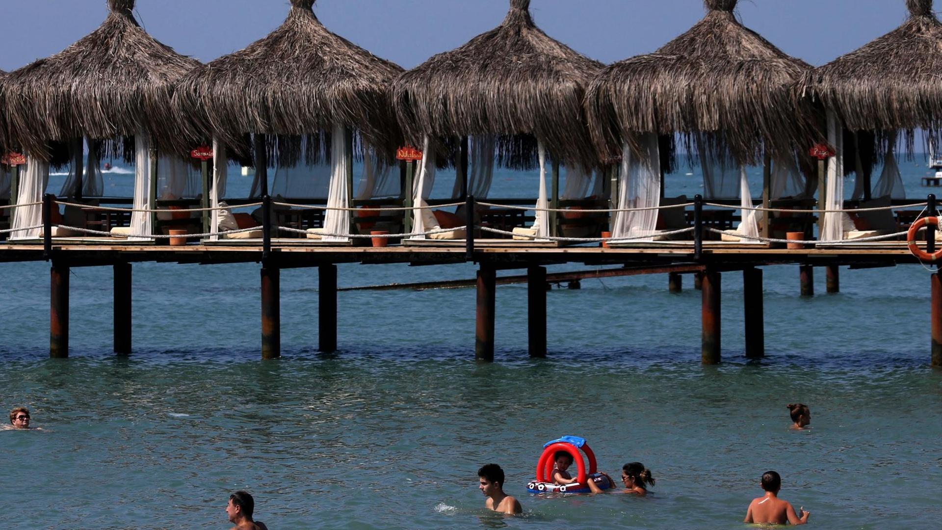 Urlaubsgäste schwimmen im Meer am Strand von Antalya. Dahinter einer Anlage mit Sonnenschirmen und Liegen, die auf Holzpfälen über dem Wasser aufgestellt ist. Urlaubsgäste schwimmen im Meer am Strand von Antalya. Dahinter einer Anlage mit Sonnenschirmen und Liegen, die auf Holzpfälen über dem Wasser aufgestellt ist.