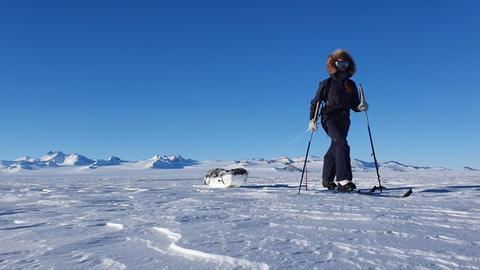 Eine Frau ist alleine auf Langlauf-Skiern mit umgeschnalltem Schlitten, auf dem ein Ruckack liegt, zu sehen.