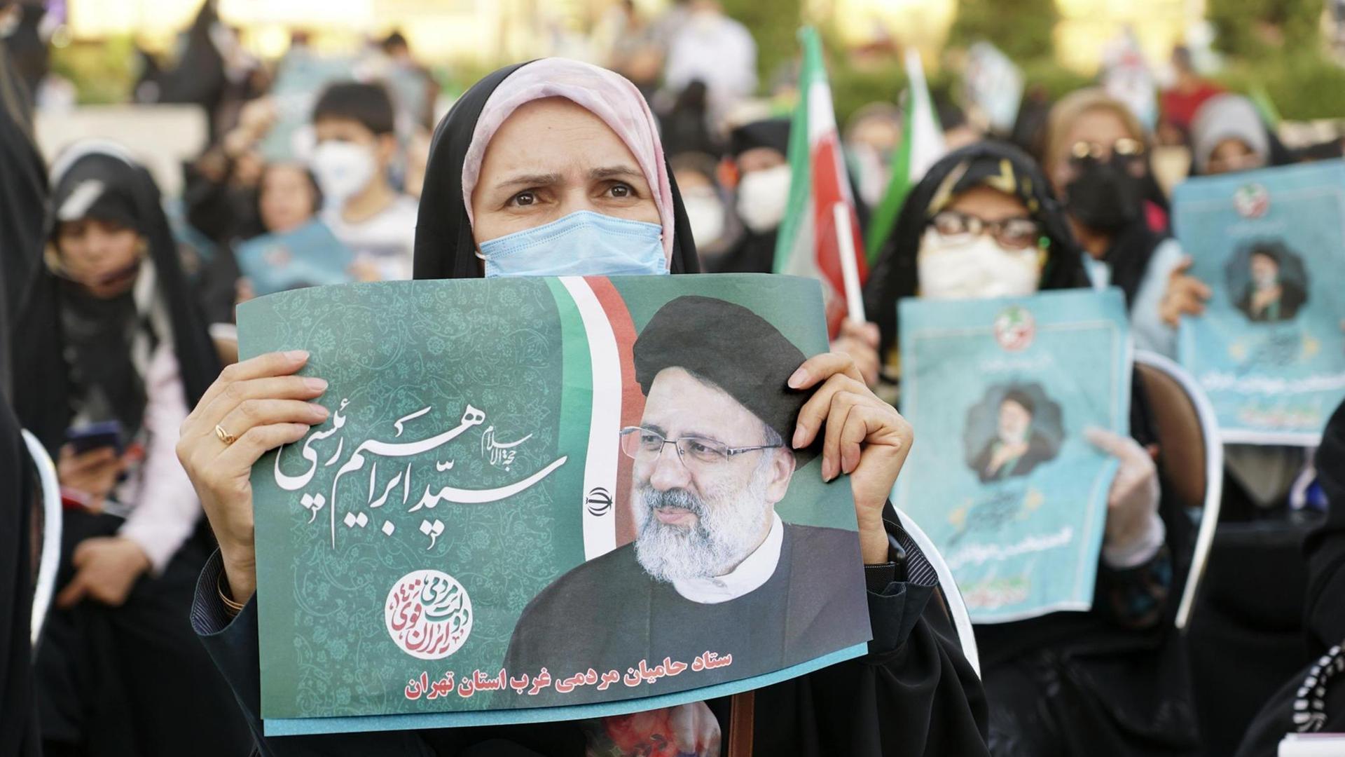 Eine Frau mit Kopftuch und Mundschutz hält im Iran ein Bild des Klerikers Ebrahim Raisi hoch. Er ist einer der Kandidaten bei der Präsidentschaftswahl.