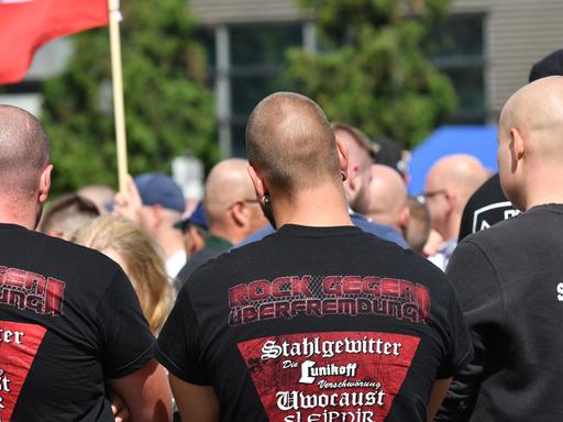 Drei Männer stehen mit dem Rücken zur Kamera, sie haben Glatzen auf dem T-Shirts des Mannes in der Mitte steht: "Rock gegen Überfremdung"