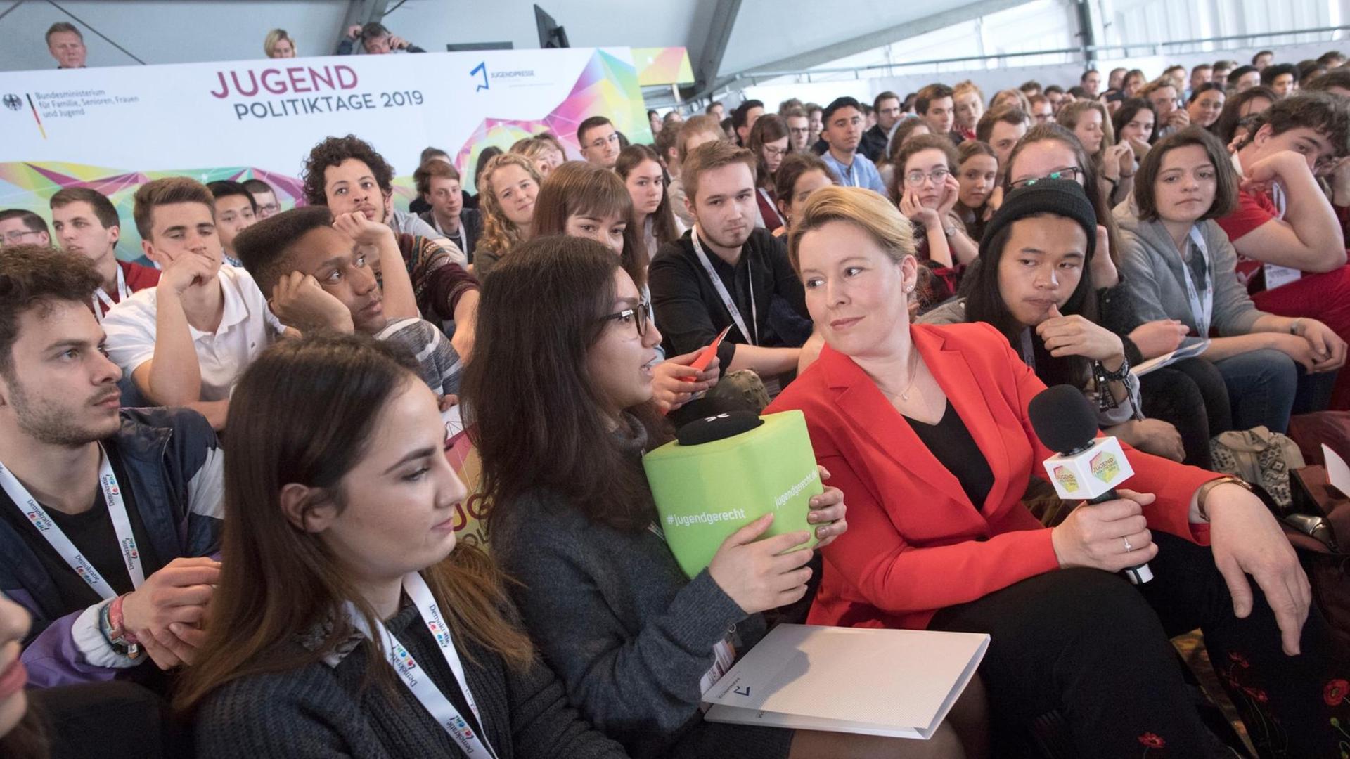 Franziska Giffey, Bundesfamilienministerin, unterhält sich bei der Eröffnung der Jugendpolitiktage 2019 mit einigen Teilnehmern. Zwischen dem 10. und 12. Mai 2019 sind rund 450 junge Menschen nach Berlin eingeladen, um über aktuelle Themen aus Politik und Gesellschaft zu diskutieren. Franziska Giffey trägt ein rotes Kleid und hält ein Mikrofon in der Hand. Franziska Giffey, Bundesfamilienministerin, unterhält sich bei der Eröffnung der Jugendpolitiktage 2019 mit einigen Teilnehmern. Zwischen dem 10. und 12. Mai 2019 sind rund 450 junge Menschen nach Berlin eingeladen, um über aktuelle Themen aus Politik und Gesellschaft zu diskutieren. Franziska Giffey trägt ein rotes Kleid und hält ein Mikrofon in der Hand.