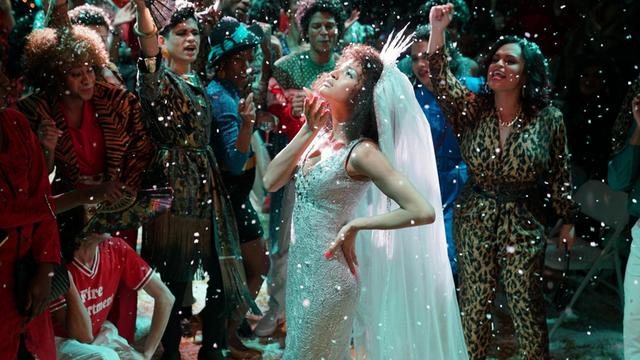 US-Schauspielerin Indya Moore steht in der Rolle der Transfrau Angel in einem weißen Glitzerkleid mit Haube umringt von anderen Tänzern auf dem Parkett, dabei regnet es Glitter