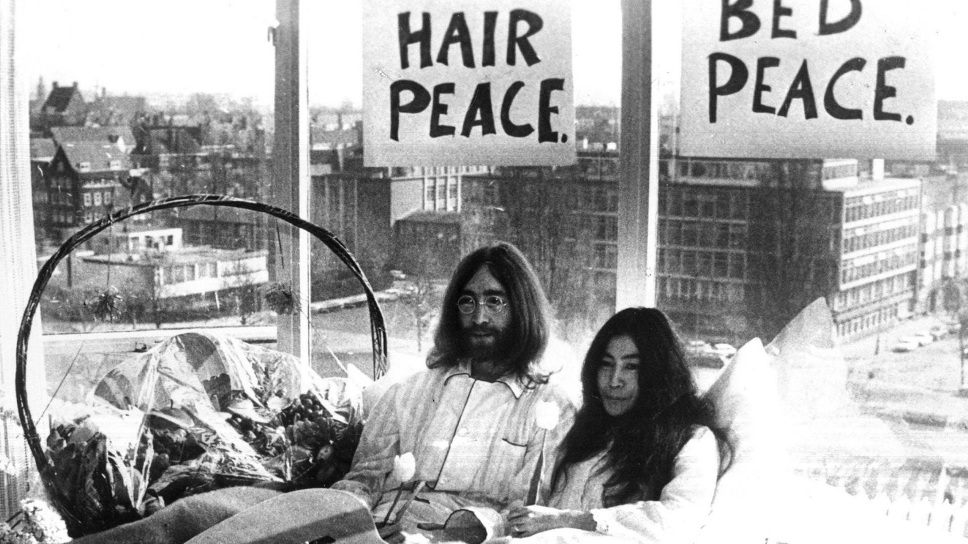 Yoko Ono und John Lennon sitzen im Bett. Über ihren Köpfen hängen Schilder mit der Aufschrift "Bed Peace" und "Hair Peace". Im Bett liegt eine Gitarre und Blumen.