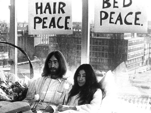 Yoko Ono und John Lennon sitzen im Bett. Über ihren Köpfen hängen Schilder mit der Aufschrift "Bed Peace" und "Hair Peace". Im Bett liegt eine Gitarre und Blumen.