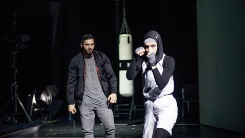 Eine Frau mit Kopftuch in Boxerpose und ein Mann stehen auf der Bühne. Im Hintergrund ist ein Boxsack zu sehen.