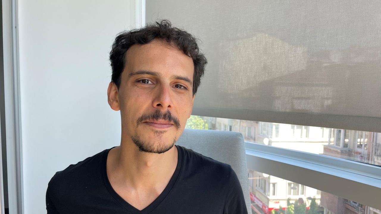 Ein junger Mann mit Bärtchen und dunklen kurzen Haaren sitzt im T-Shirt am Fenster. Ein junger Mann mit Bärtchen und dunklen kurzen Haaren sitzt im T-Shirt am Fenster.