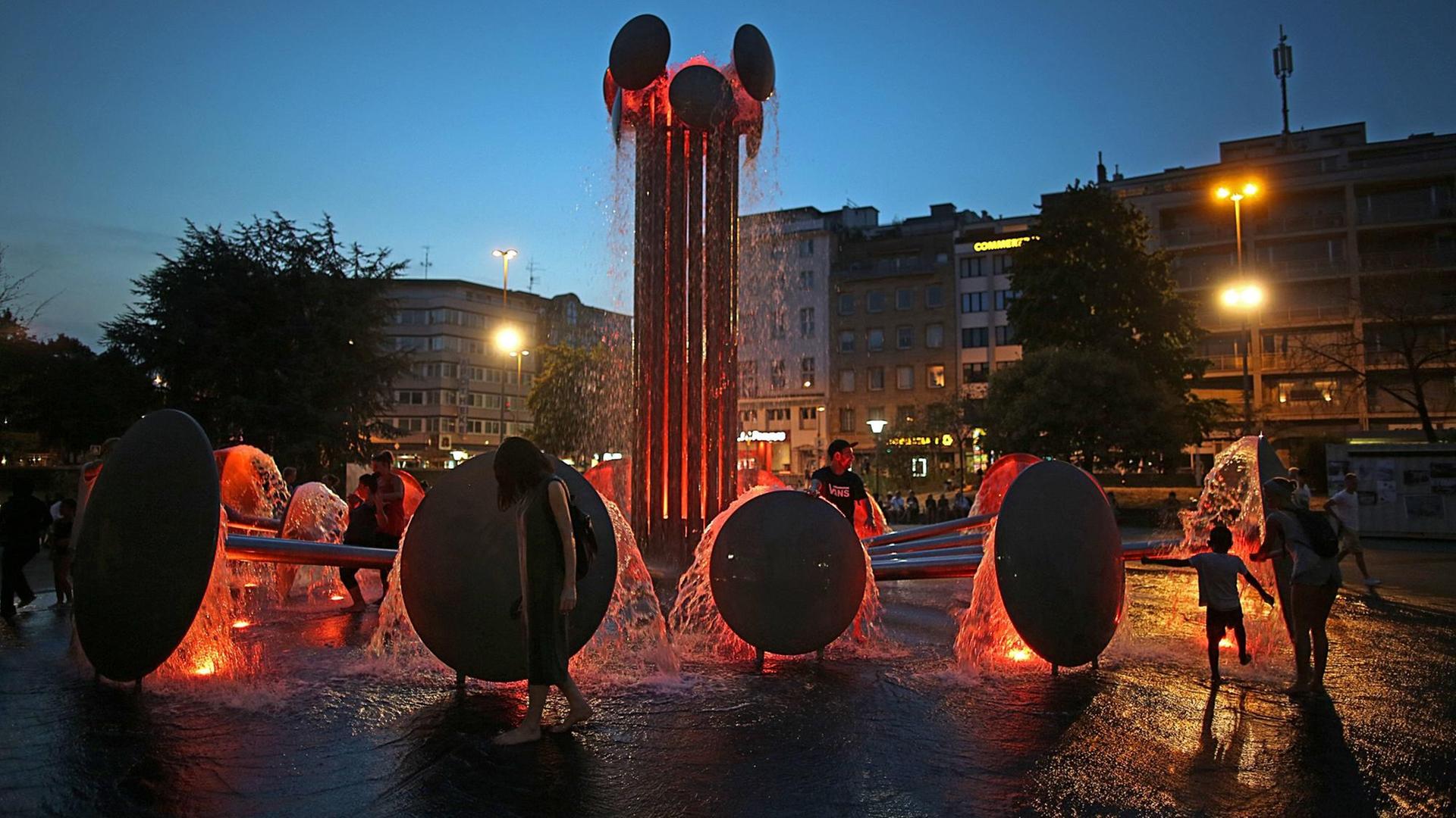 Menschen erfrischen sich am Abend am rot beleuchteten Brunnen am Kölner Ebertplatz.