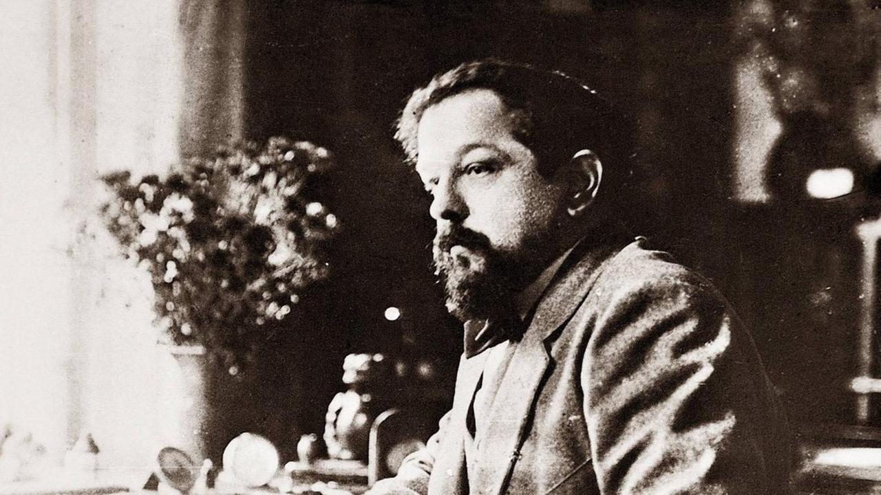Schwarz-Weiß-Aufnahme von Claude Debussy um 1918. Der Komponist sitzt an einem Tisch und hat seinen Ellenbogen auf die polierte Holzplatte gestützt.