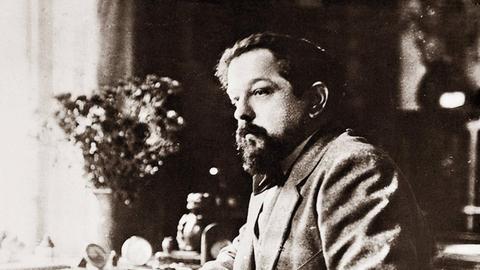 Schwarz-Weiß-Aufnahme von Claude Debussy um 1918. Der Komponist sitzt an einem Tisch und hat seinen Ellenbogen auf die polierte Holzplatte gestützt