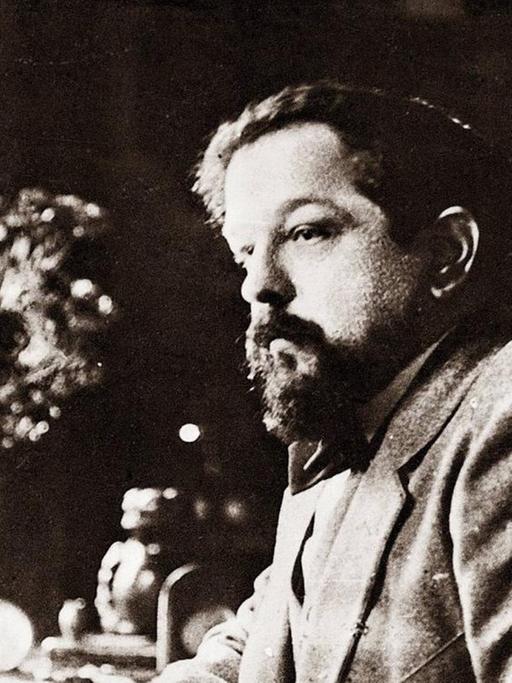 Schwarz-Weiß-Aufnahme von Claude Debussy um 1918. Der Komponist sitzt an einem Tisch und hat seinen Ellenbogen auf die polierte Holzplatte gestützt