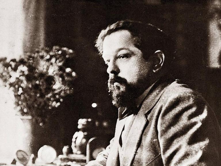 Schwarz-Weiß-Aufnahme von Claude Debussy um 1918. Der Komponist sitzt an einem Tisch und hat seinen Ellenbogen auf die polierte Holzplatte gestützt