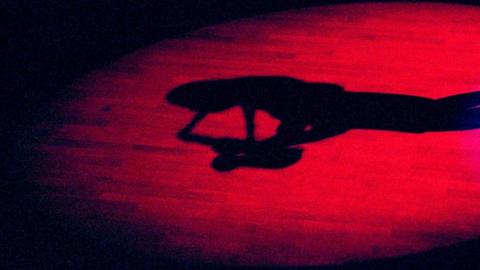 Ein roter Lichtkegel auf einem Holzboden zeigt den Schatten einer Saxofon spielenden Frau.