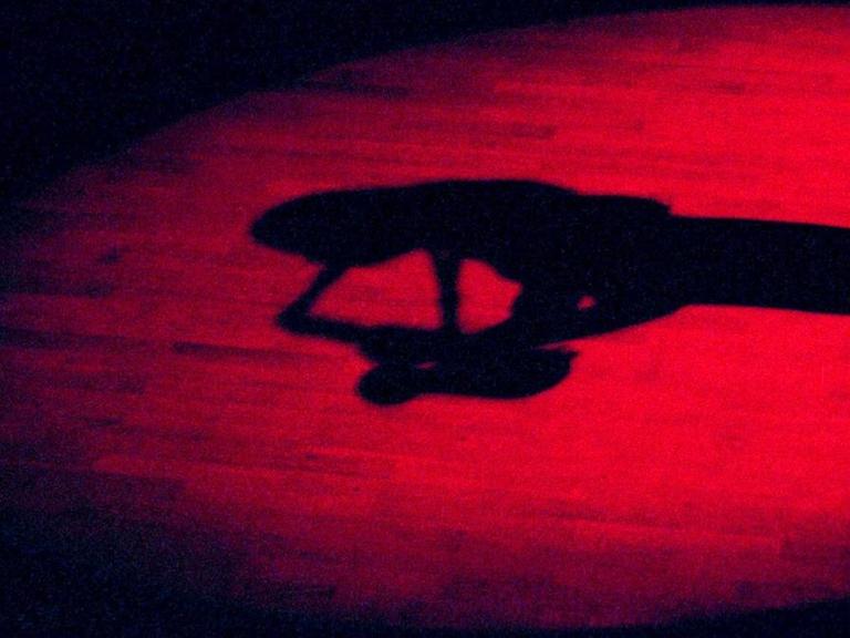 Ein roter Lichtkegel auf einem Holzboden zeigt den Schatten einer Saxofon spielenden Frau.