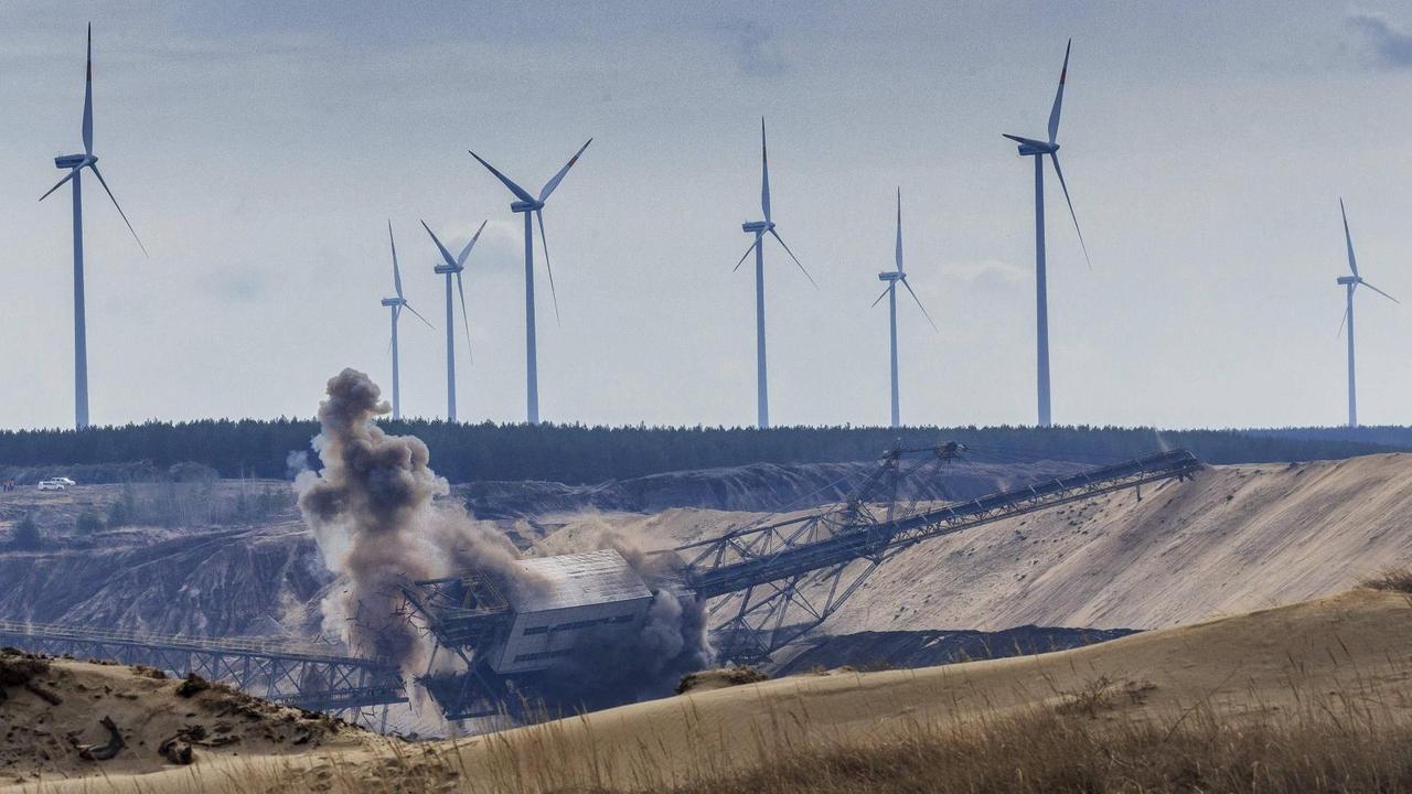 Panorama eines Bergbaugebiets mit einer Abbau-Brücke, die gerade gesprengt wird. Im Hintergrund des Bild stehen hohe Windkraftanlagen und bilden dadurch zum Vordergrund einen energiepolitischen Kontrast.