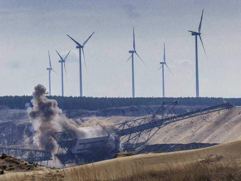 Panorama eines Bergbaugebiets mit einer Abbau-Brücke, die gerade gesprengt wird. Im Hintergrund des Bild stehen hohe Windkraftanlagen und bilden dadurch zum Vordergrund einen energiepolitischen Kontrast.