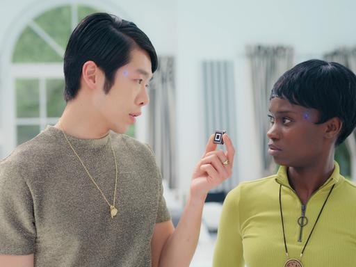 Cee (Lorna Ishema) und Kim (Yung Ngo) stehen sich schräg gegenüber. Kim hält einen kleinen quadratischen Gegenstand vor Cees Gesicht, die ungläubig blickt. An ihren Schläfen leuchten blaue Leuchtdioden.