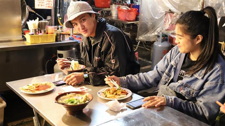 Ein asiatischer Mann und eine asiatische Frau essen auf dem Nachtmarkt in Taipeh mit Stäbchen Ein asiatischer Mann und eine asiatische Frau essen auf dem Nachtmarkt in Taipeh mit Stäbchen