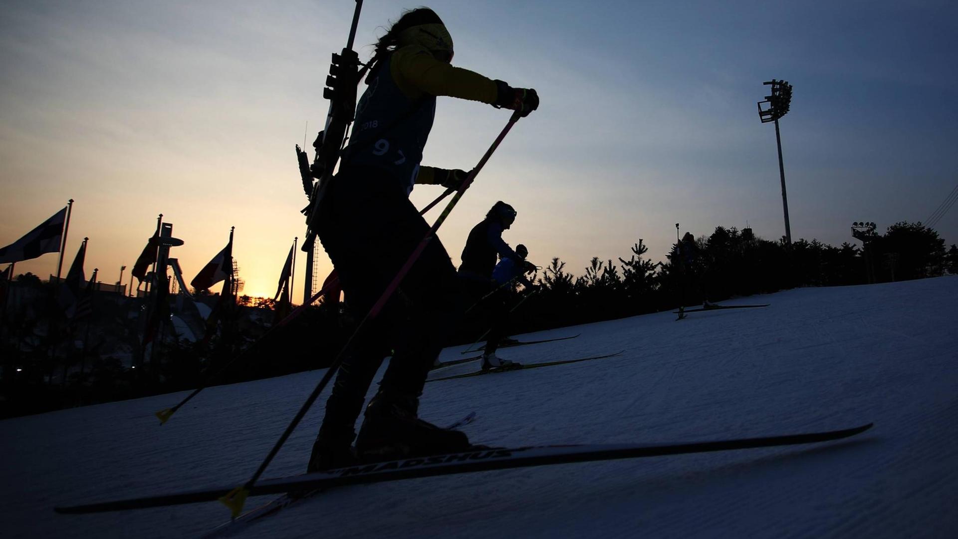 Eine Biathletin fährt im Gegenlicht der untergehenden Sonne über die Piste, im Hintergrund sind weitere Sportlerinnen zu erkennen. Eine Biathletin fährt im Gegenlicht der untergehenden Sonne über die Piste, im Hintergrund sind weitere Sportlerinnen zu erkennen.