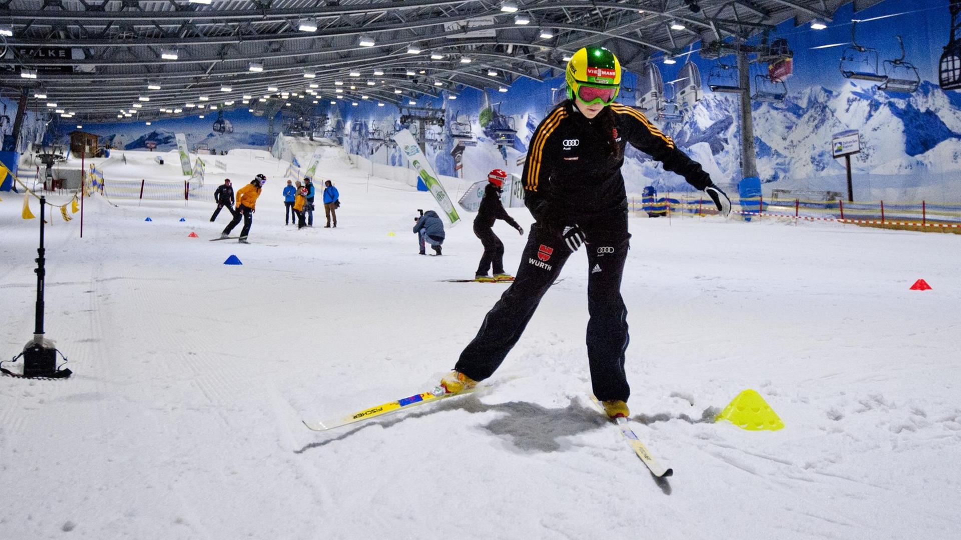 Eine Skispringerin fährt auf einer Piste, die sich in einer Halle befindet. Im Hintergrund sind weitere Skifahrerinnen zu sehen. Auf den Wänden sind Berge gemalt. Eine Skispringerin fährt auf einer Piste, die sich in einer Halle befindet. Im Hintergrund sind weitere Skifahrerinnen zu sehen. Auf den Wänden sind Berge gemalt.