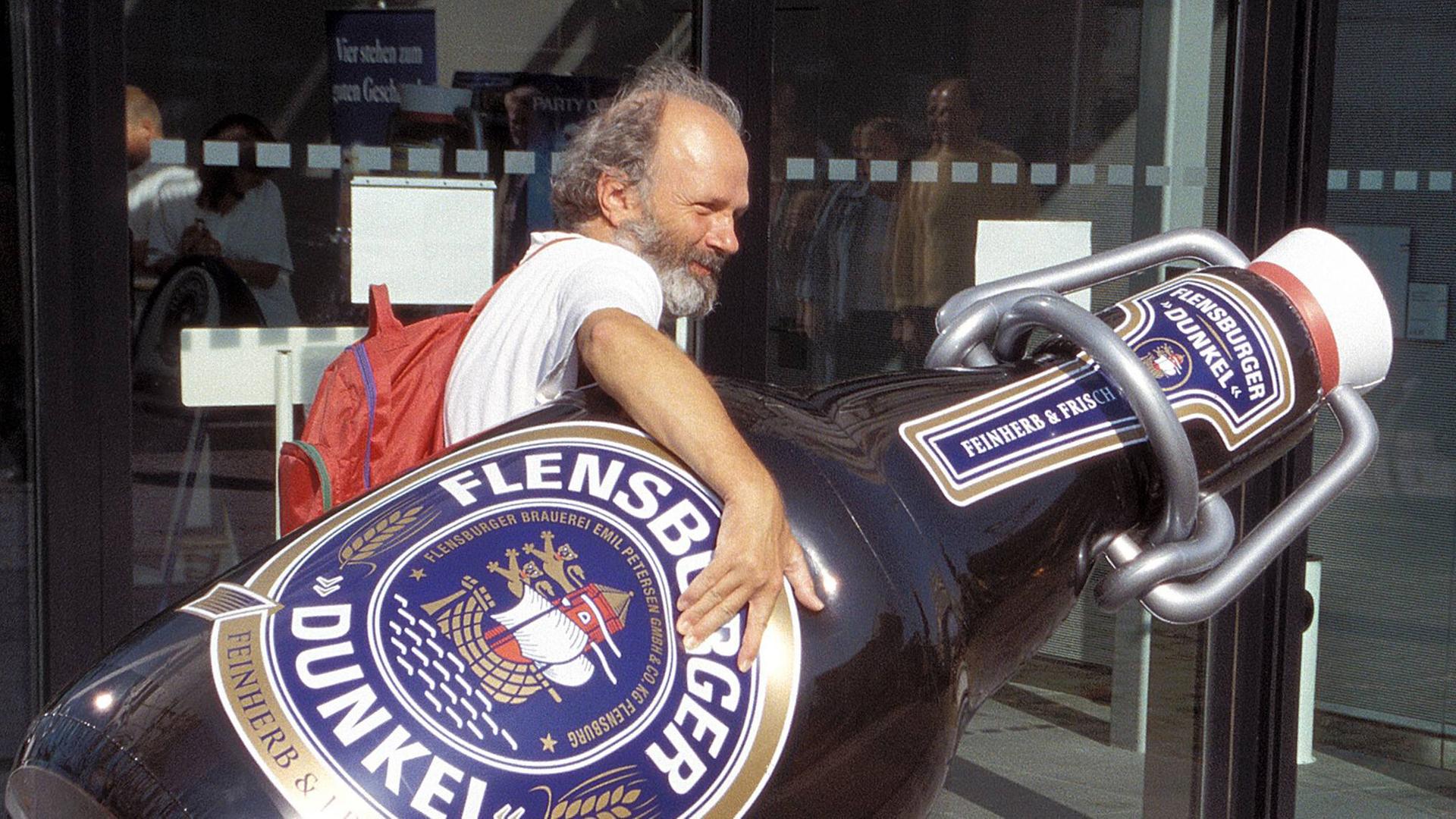 Ein bärtiger Mann trägt eine überdimensionale Flensburger-Bierflasche Ein bärtiger Mann trägt eine überdimensionale Flensburger-Bierflasche