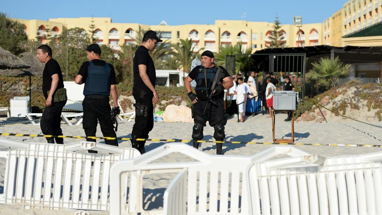 Vier schwarzgekleidete Polizisten stehen einen Tag nach dem Attentat am Strand vor dem Hotel Imperial Marhaba: Die Polizisten tragen Schusswesten. Im Vordergrund sieht man umgestürzte Liegestühle.