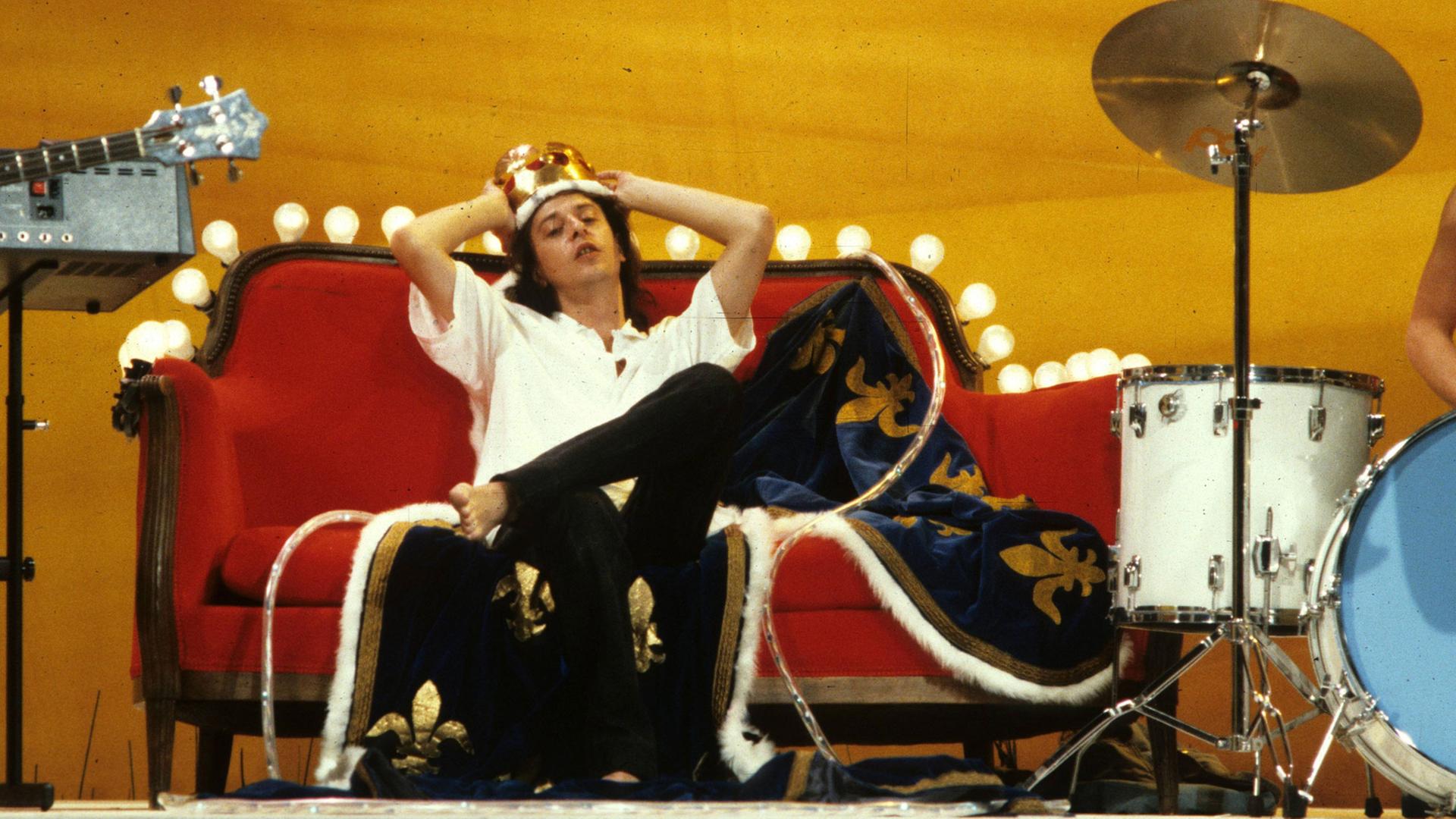 Der Sänger Rio Reiser liegt auf einem roten Sofa, dessen Lehne mit einer Lichterkette geschmückt ist und trägt eine goldete Krone aus Plastik.