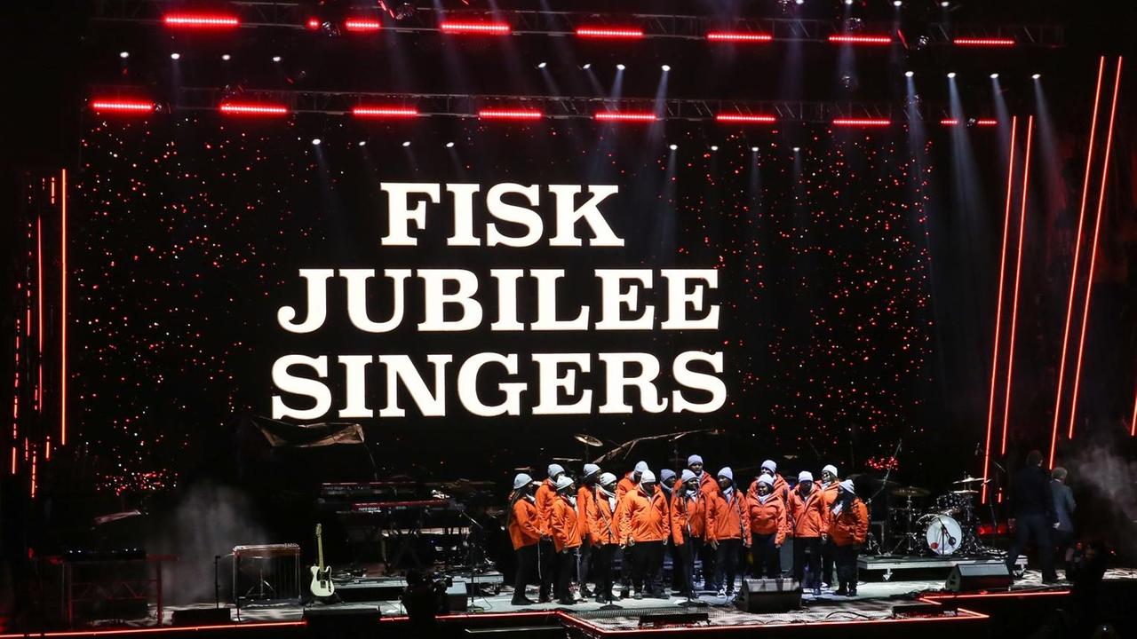 Auf einer großen Bühne, auf deren Hintergrund der Name des Chores in Großbuchstaben eingeblendet wird, steht der Chor geschlossen mit roten Shirts und singt.