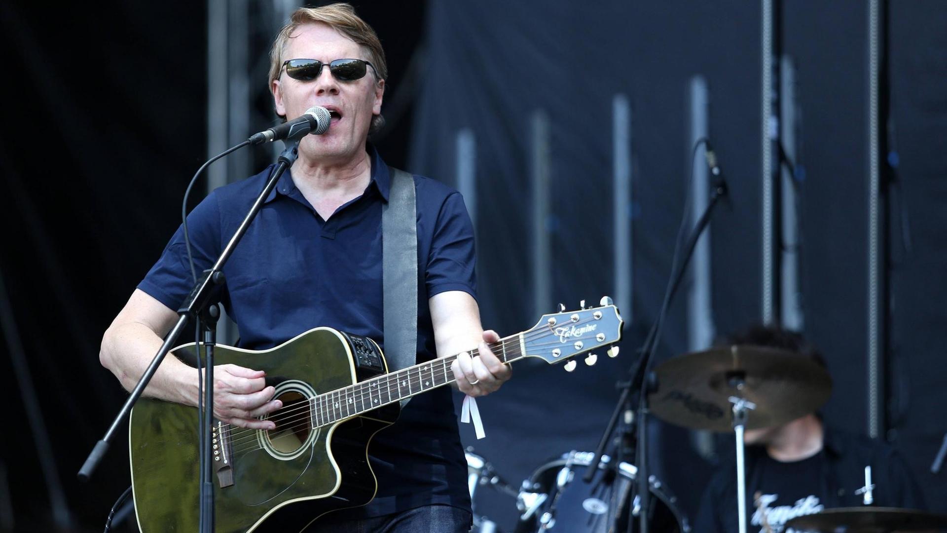 Chris Eckman steht mit Sonnenbrille und Gitarre auf der Bühne. Im Hintergrund ist ein Schlagzeug zu sehen. Chris Eckman steht mit Sonnenbrille und Gitarre auf der Bühne. Im Hintergrund ist ein Schlagzeug zu sehen.