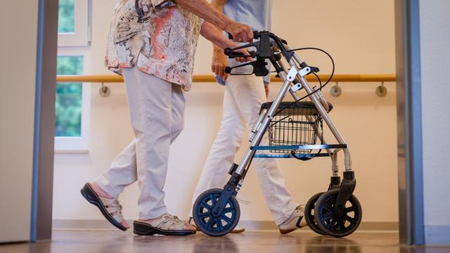 Eine Altenpflegerin begleitet eine ältere Frau mit dem Rollator durch den Flur eines Pflegeheims.