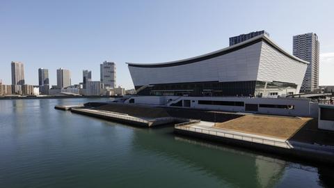 Blick auf eine Wasserfläche an deren Ufer eine moderne Sportarena steht — im Hintegrund sind Hochhäuser zu sehen