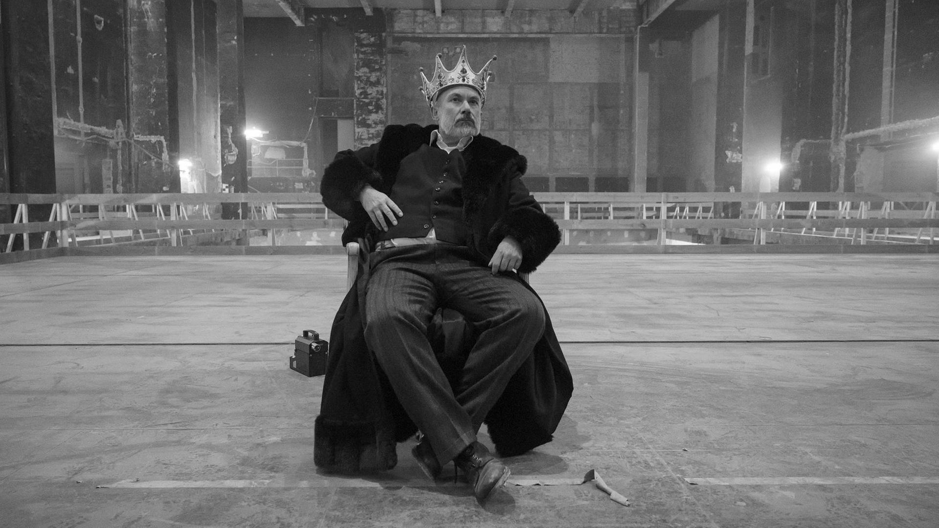 Der Schauspieler Klaus Müller sitzt mit einer Krone auf einer riesigen Bühne