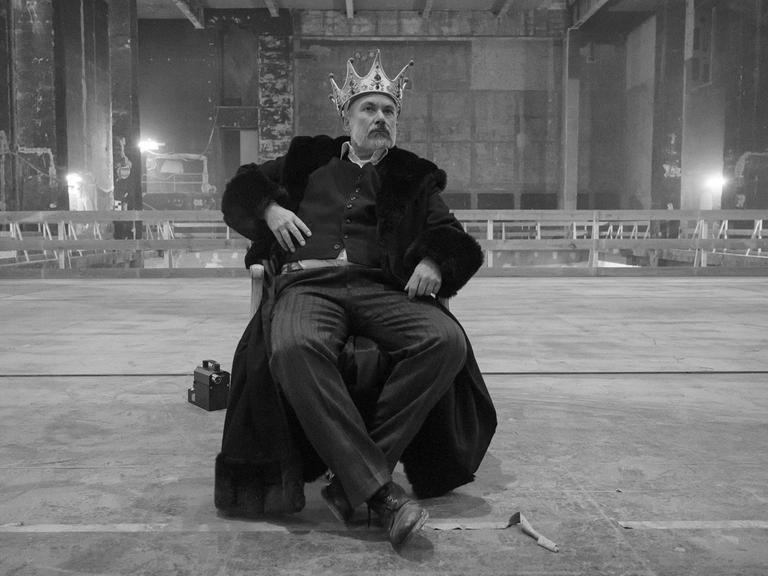 Der Schauspieler Klaus Müller sitzt mit einer Krone auf einer riesigen Bühne