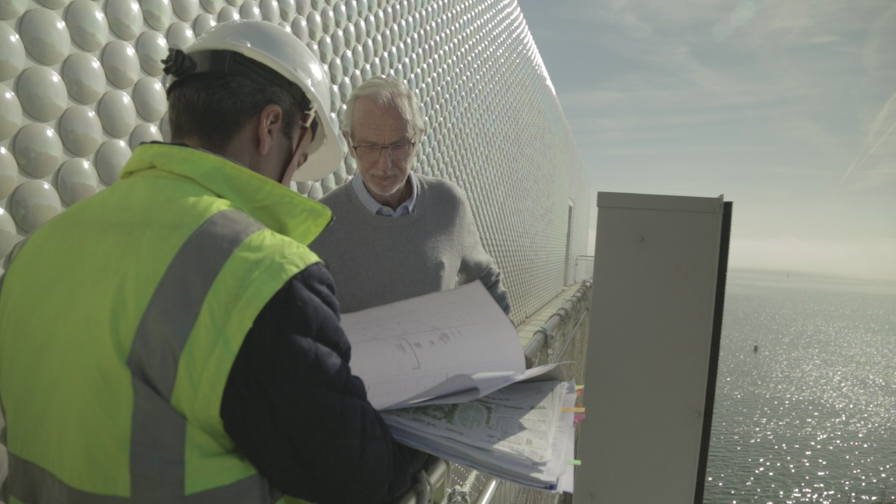Auf dem Bild sieht man den Architekten Renzo Piano mit einem Mann in Schutzhelm auf der Baustelle. Sie schauen gemeinsam auf Pläne.