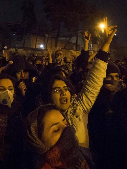 Iranische Studenten demonstrieren in Teheran. Es ist dunkel, Frauen und Männer halten ihre Hände in die Höhe und rufen, ein Plakat ist zu sehen.