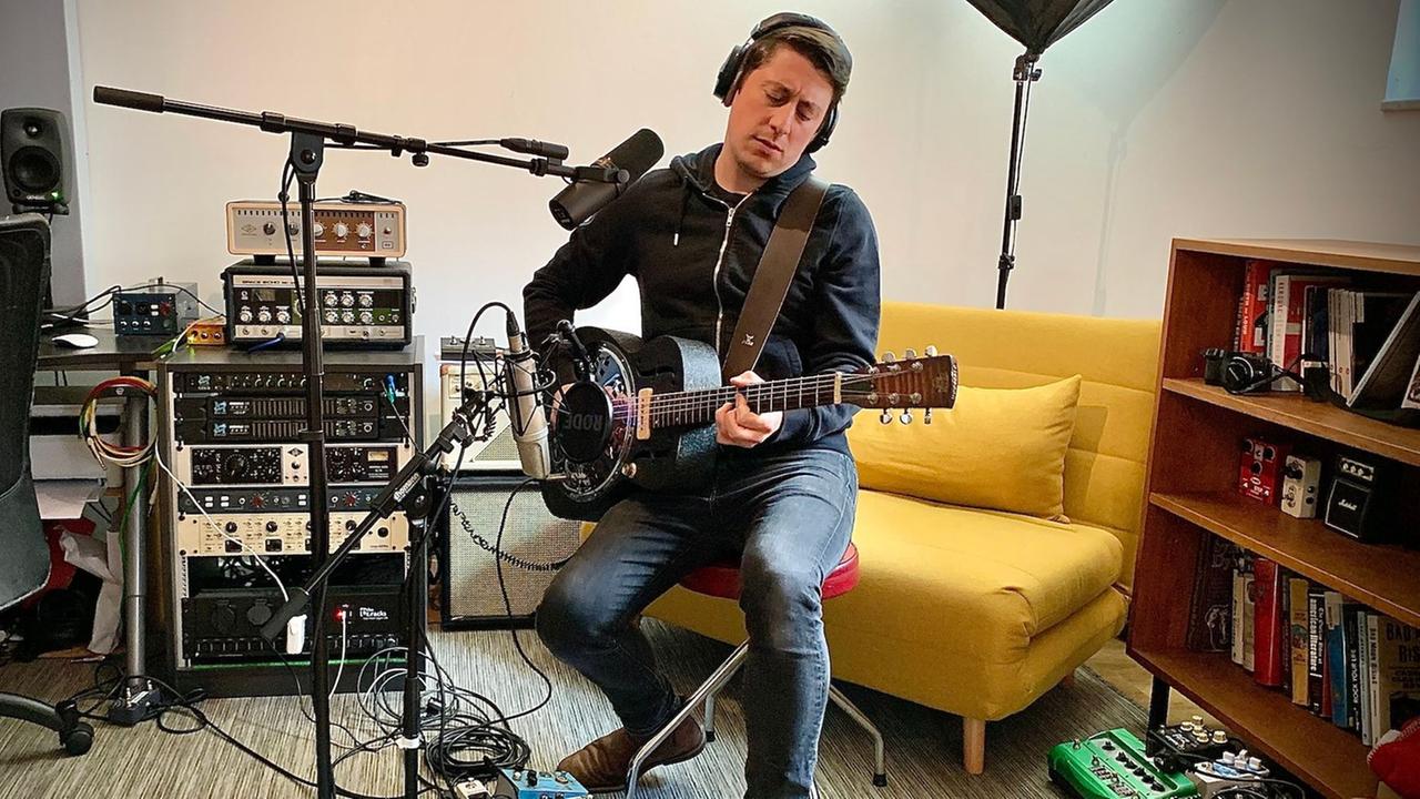 Ein Mann mit schwarzen kurzen Haaren sitzt auf einem Hocker vor Mikrofonen und spielt Gitarre. Er trägt Kopfhörer, hinter ihm steht ein gelber Sessel, ein Regal und technisches Equipment. Ein Mann mit schwarzen kurzen Haaren sitzt auf einem Hocker vor Mikrofonen und spielt Gitarre. Er trägt Kopfhörer, hinter ihm steht ein gelber Sessel, ein Regal und technisches Equipment.