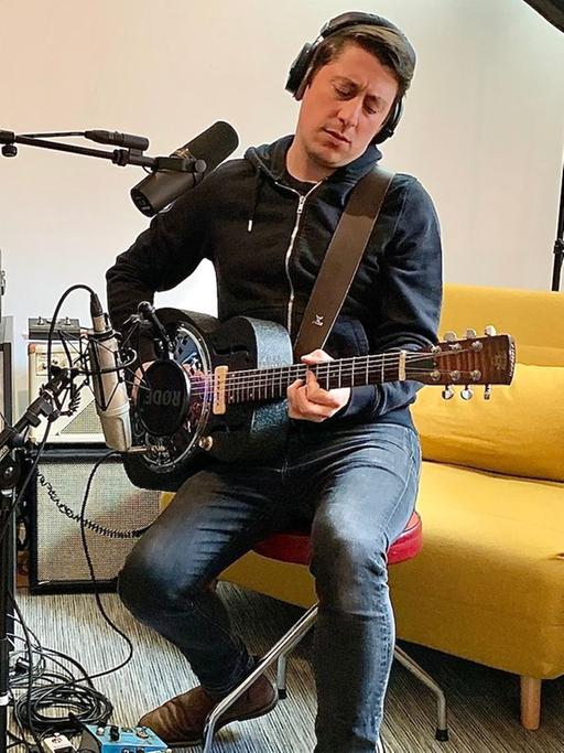 Ein Mann mit schwarzen kurzen Haaren sitzt auf einem Hocker vor Mikrofonen und spielt Gitarre. Er trägt Kopfhörer, hinter ihm steht ein gelber Sessel, ein Regal und technisches Equipment.
