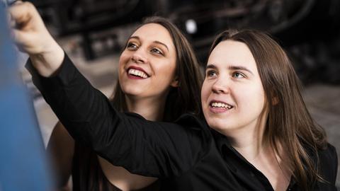 Die zwei Musikerinnen stehen lachend vor einer Wand und blicken bzw. zeigen nach oben auf etwas nicht Erkennbares