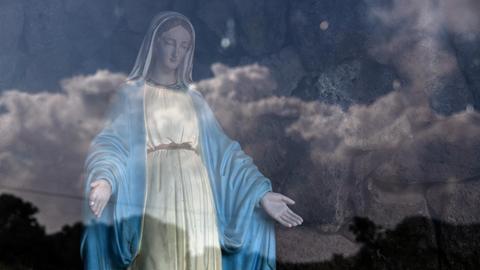 Eine Jungfrau Maria Statue hinter Glas, in dem sich der Himmel spiegelt.