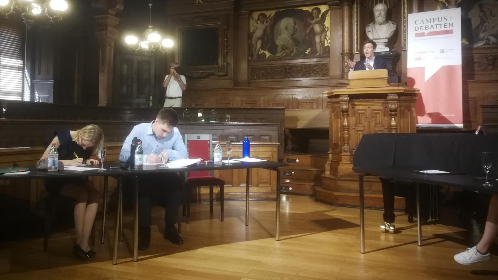 Eine Frau und ein Mann, Finalisten der Debattiermeisterschaft, sitzen an einem Tisch und bereiten sich vor, ein Mann debattiert. Eine Frau und ein Mann, Finalisten der Debattiermeisterschaft, sitzen an einem Tisch und bereiten sich vor, ein Mann debattiert.