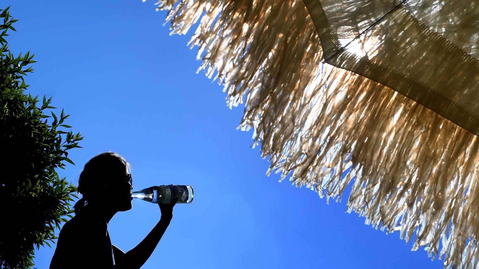 Eine Frau steht unter einem Sonnenschirm und trinkt aus einer Wasserflasche 