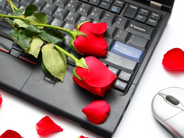 Warum werden so viele Menschen Opfer von Romance Scams? Zu sehen: Eine Tastatur auf einem Tisch, rote Rosen darauf und eine Maus.