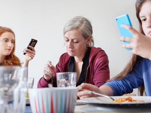 Eine Mutter isst mit ihren Töchtern zu Mittag, während die Töchter auf ihre Smartphones schauen.