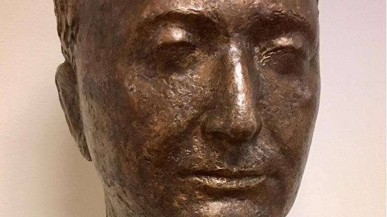 Eine Bronzestatue zeigt den Kopf eines Mannes mit hoher Stirn und sanftem Lächeln.
