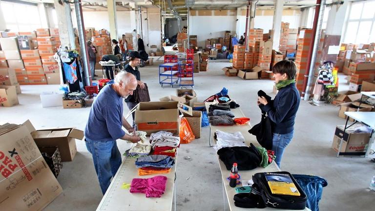 Man sieht eine große Halle mit Kartons voller Kleidung, an Tischen stehen Menschen und sortieren die Spenden. Man sieht eine große Halle mit Kartons voller Kleidung, an Tischen stehen Menschen und sortieren die Spenden.