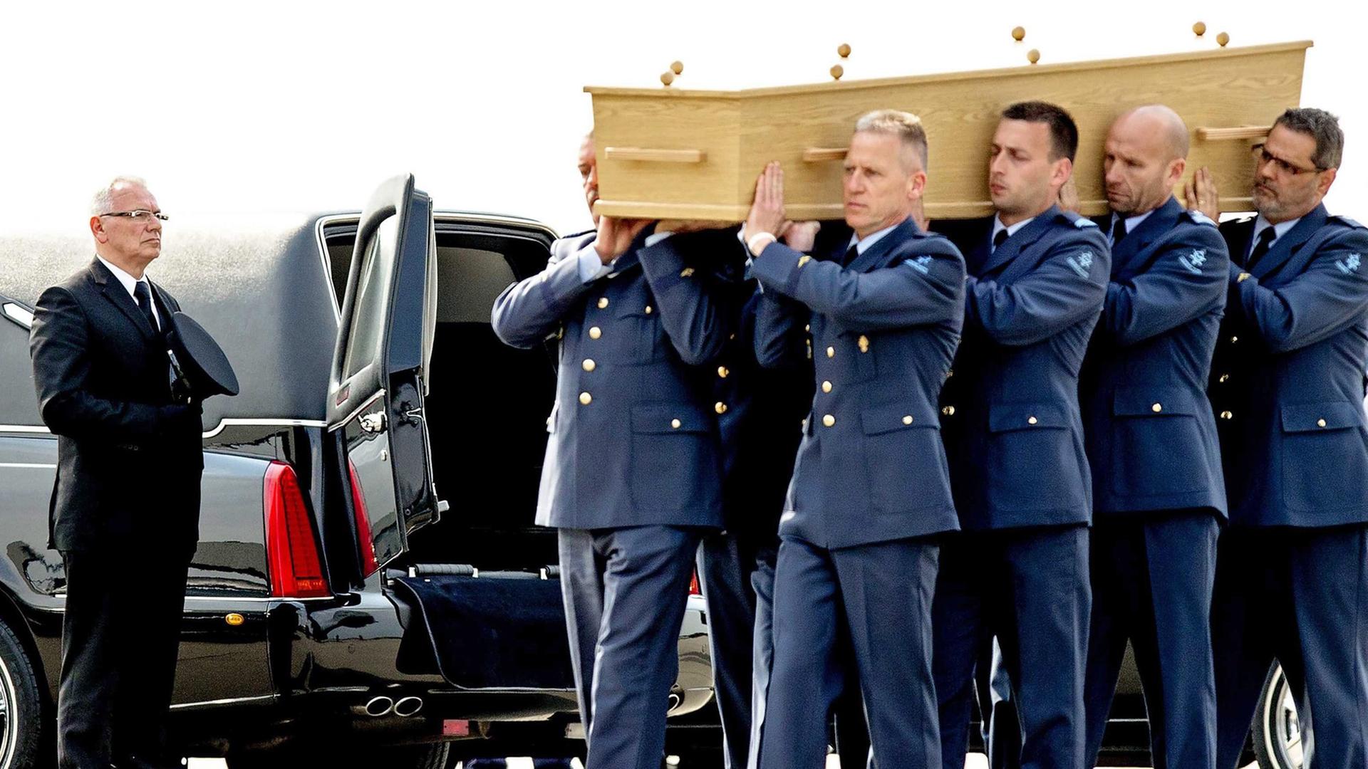 Acht Militärangehörige in blauen Uniformen tragen einen Holzsarg auf ihren Schultern, im Hintergrund das schwarze Fahrzeug eines Bestatters mit geöffneten Hecktüren.