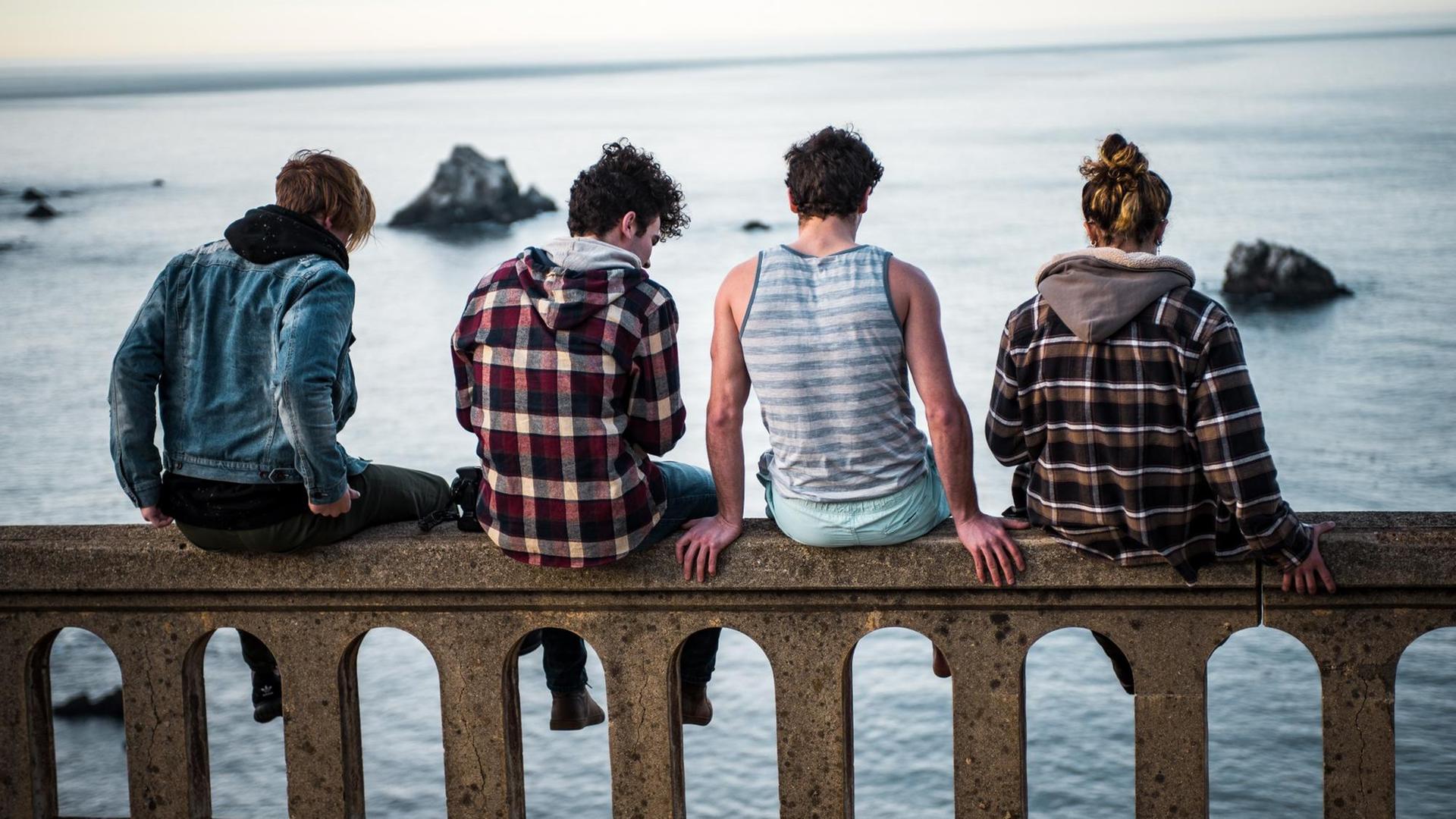 Drei Jungs und ein Mädchen sitzen mit dem Rücken zur Kamera auf einer Mauer und schauen aufs Meer. Drei Jungs und ein Mädchen sitzen mit dem Rücken zur Kamera auf einer Mauer und schauen aufs Meer.