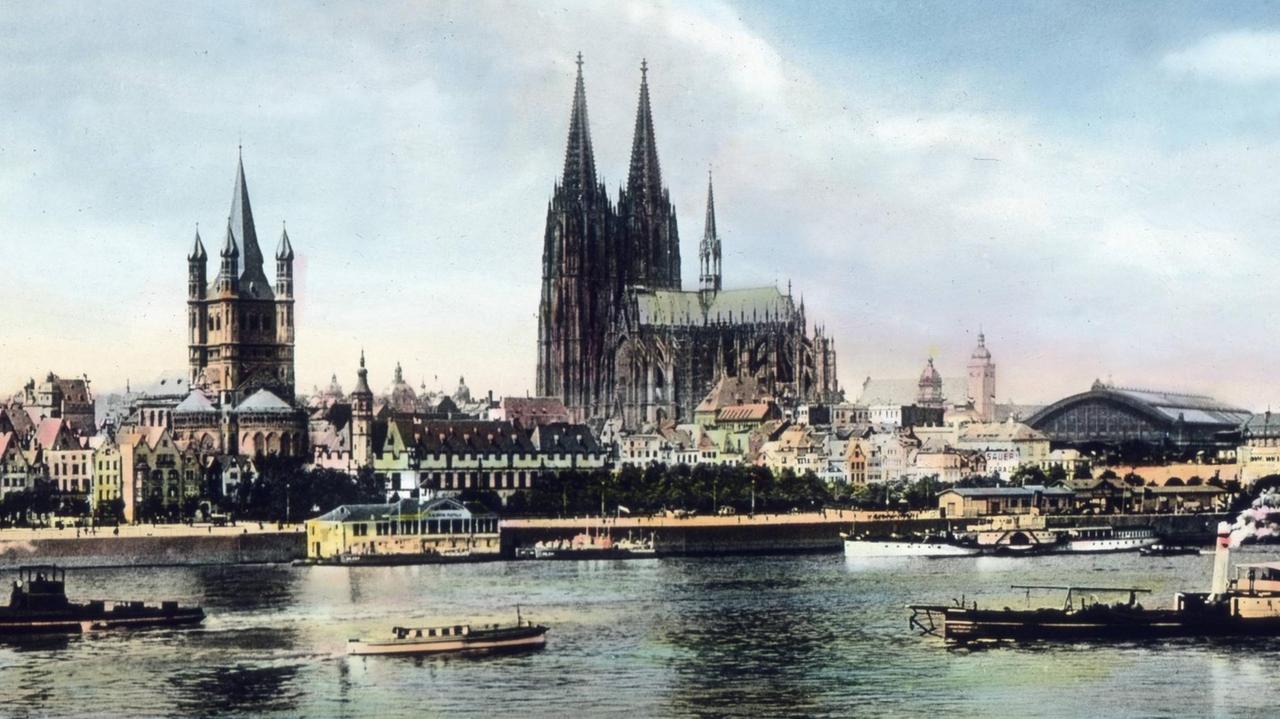 Eine nachkolorierte Stadtansicht vom Rheinufer der Stadt Köln mit fertig erstelltem Dom und viele historische Gebäude am Ufer samt alten Dampfschiffen auf dem Fluss.