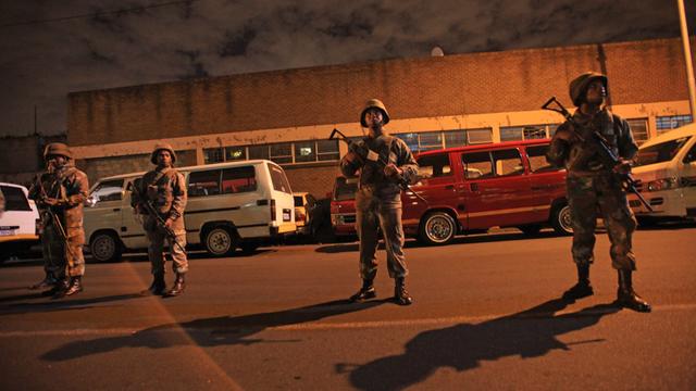 Vier bewaffnete Soldaten stehen bei Nacht auf einer Straße, im Hintergrund mehrere Autos.